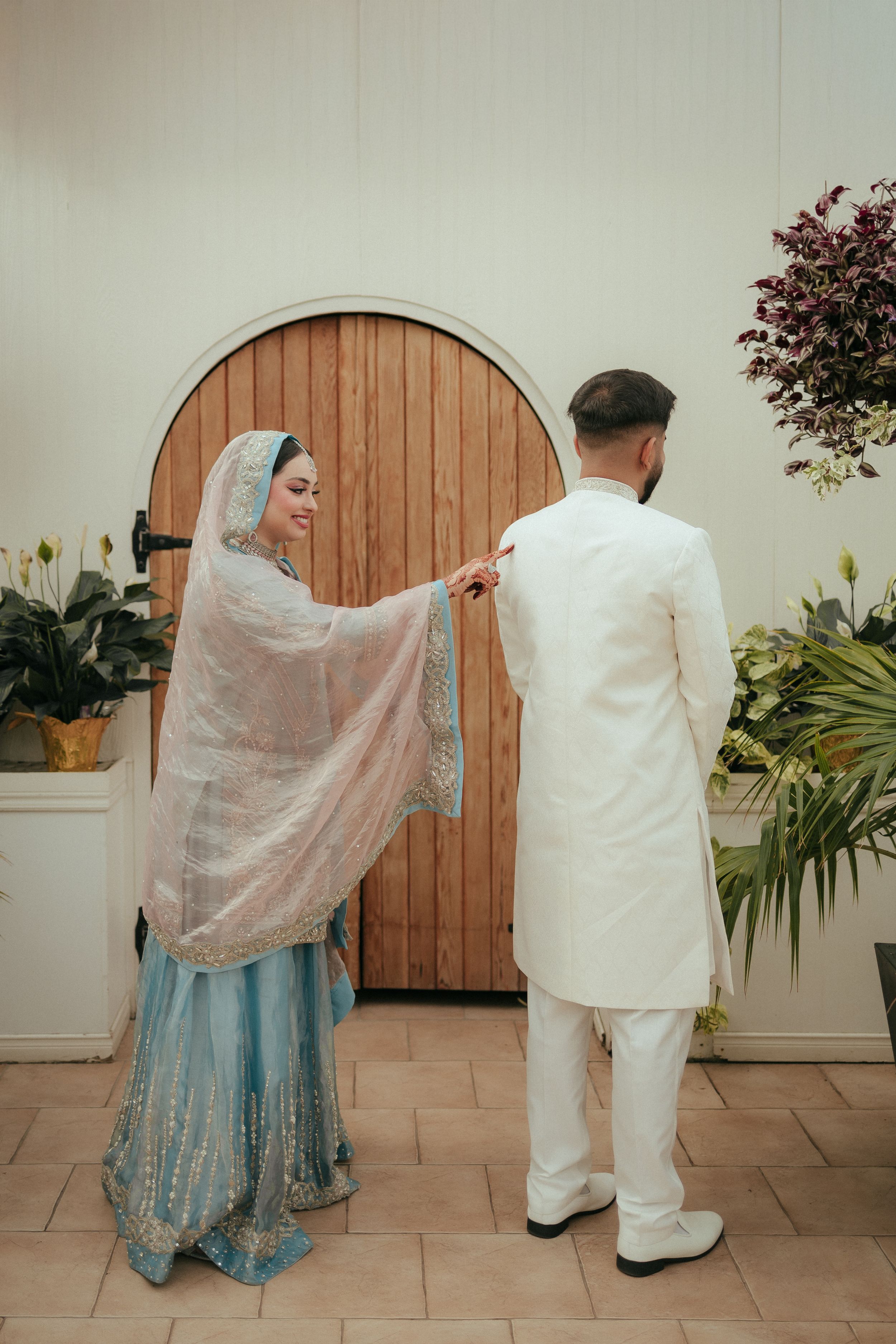 Traditional wedding couple in white and blue attire stand before wooden arched door in greenhouse.