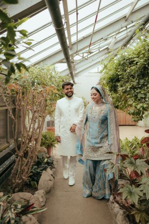 Wedding couple walks hand in hand through lush greenhouse pathway in traditional attire.