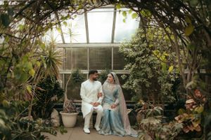 Couple in traditional wedding attire pose together in greenhouse surrounded by lush plants.