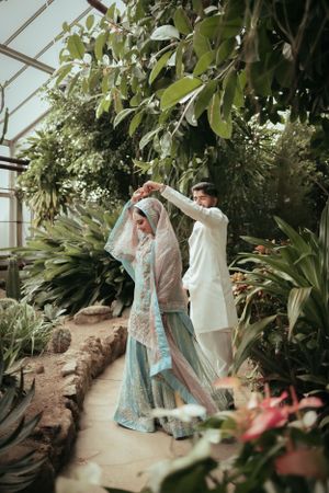 Wedding couple in white and blue traditional attire dance together in greenhouse setting.