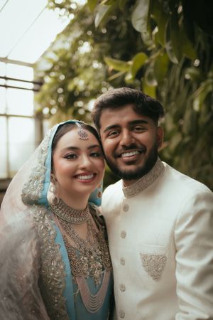 Newlyweds in traditional South Asian wedding attire share joyful moment in greenhouse.