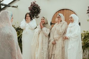 Group in white traditional garments gather for wedding celebration in greenhouse setting.