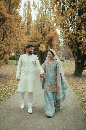 A couple walks together in autumn scenery wearing traditional South Asian wedding attire.