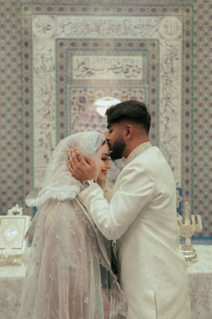 A tender moment between a couple in front of decorative Islamic calligraphy.