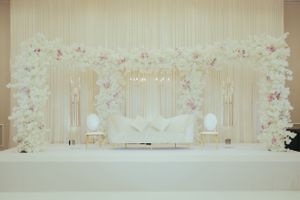 Elegant wedding stage backdrop with white floral arch and pink accents decorating a luxurious seating area.