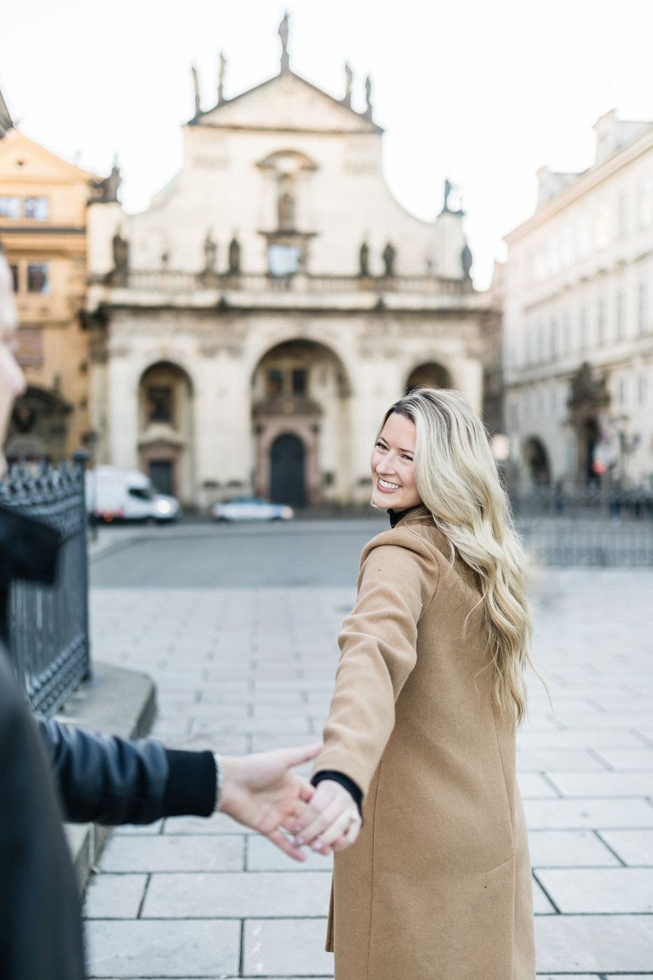 Woman in camel coat leading someone by hand near historic European church building.