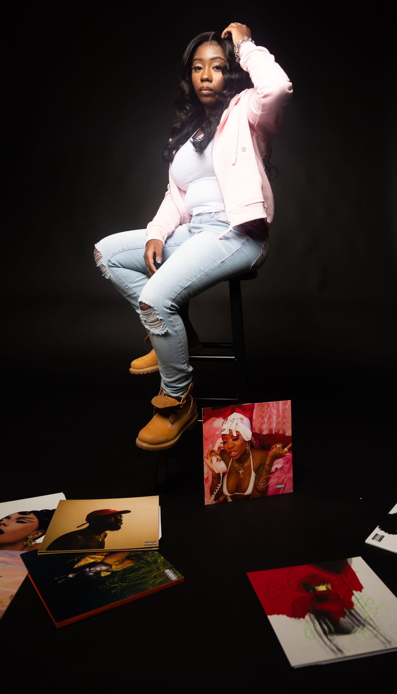Young woman in pink jacket and light jeans sits on stool with scattered photo books on dark background.