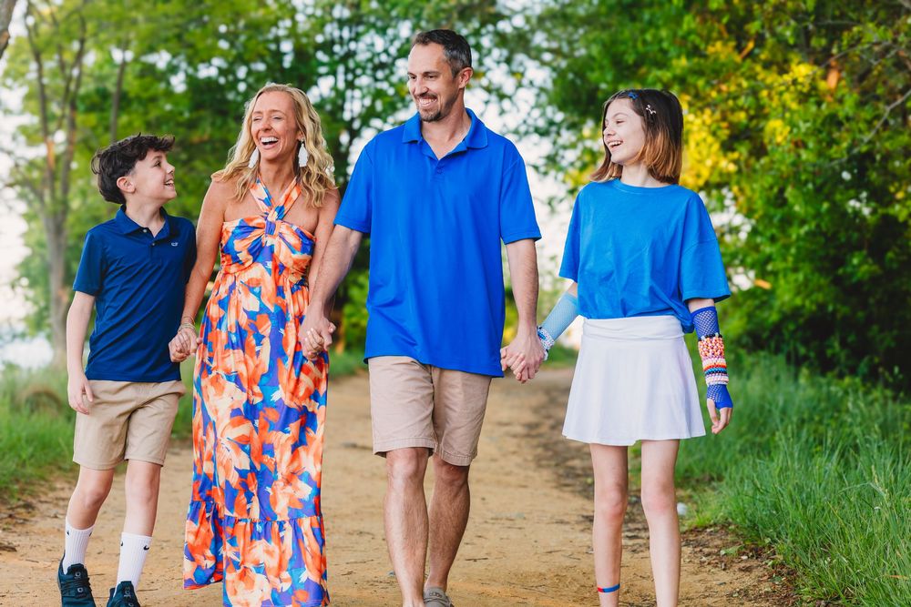 Happy family of four walking hand-in-hand on a scenic dirt path surrounded by lush green trees.