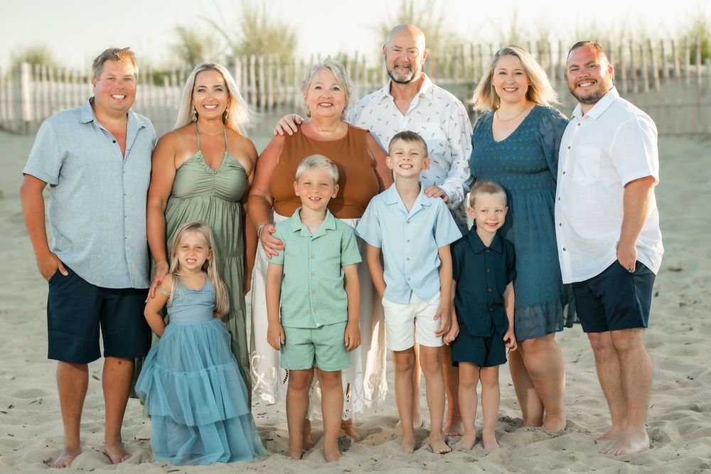 Family of ten posing together on sandy beach, smiling in coordinated blue and green outfits with white fence backdrop.