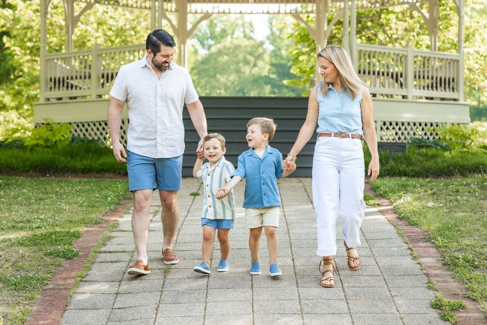 Family of four walking hand-in-hand on a brick path in a sunny park near a white gazebo.