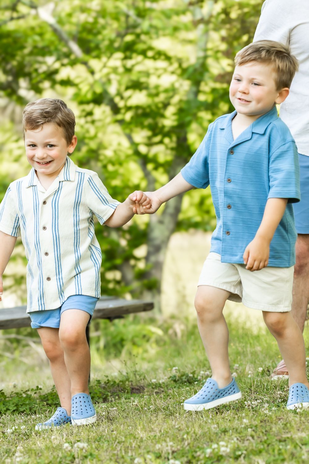 Two young boys in blue outfits hold hands while walking on green grass in a sunny park.