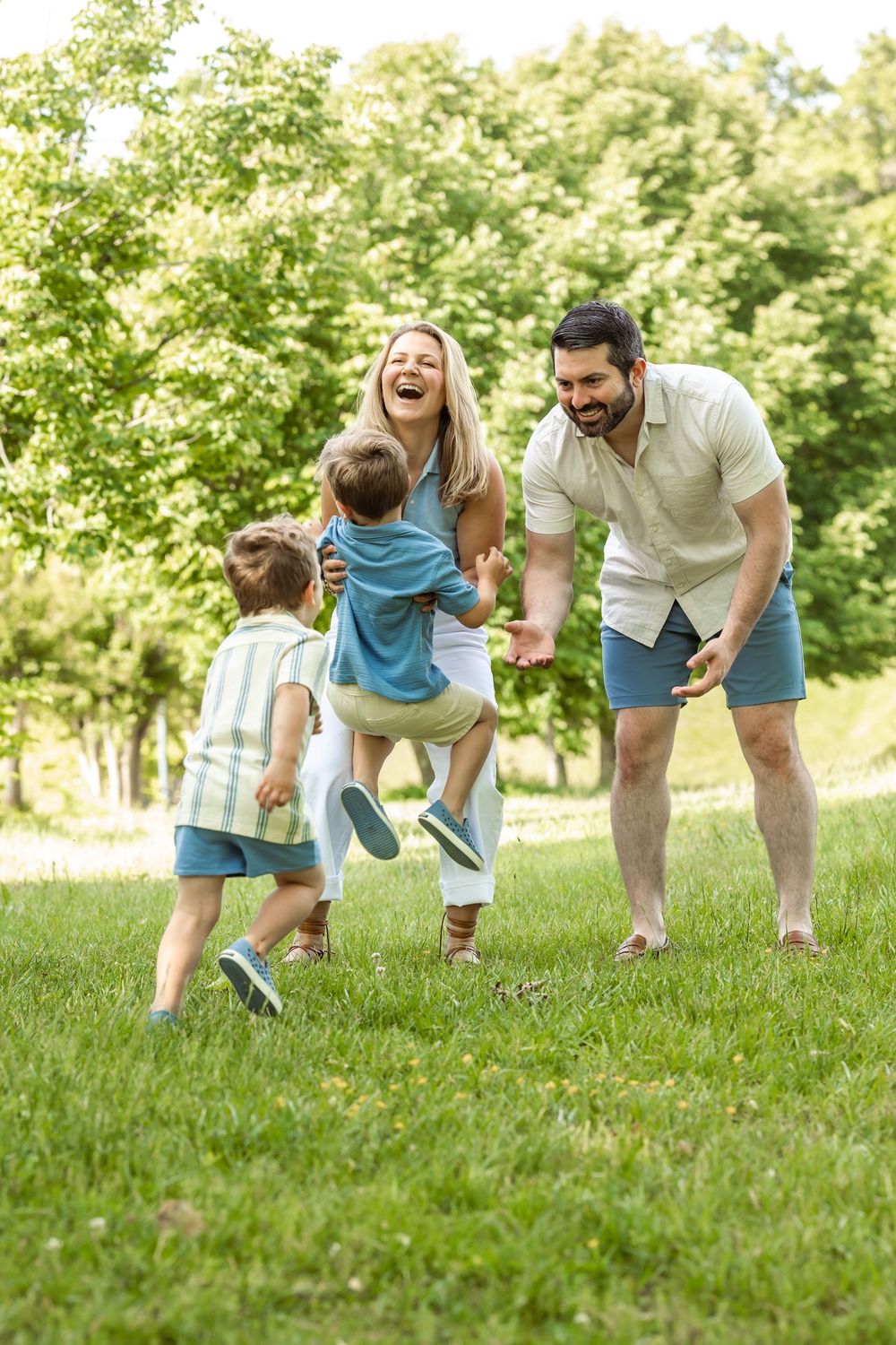 Happy family with two young boys playing and laughing together on green grass in a sunny park.