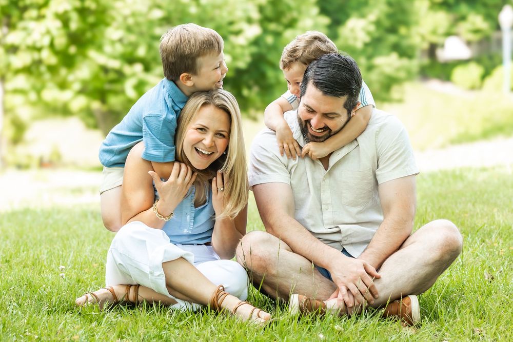 Happy family of four sitting on green grass, two children hugging parents from behind on a sunny day.