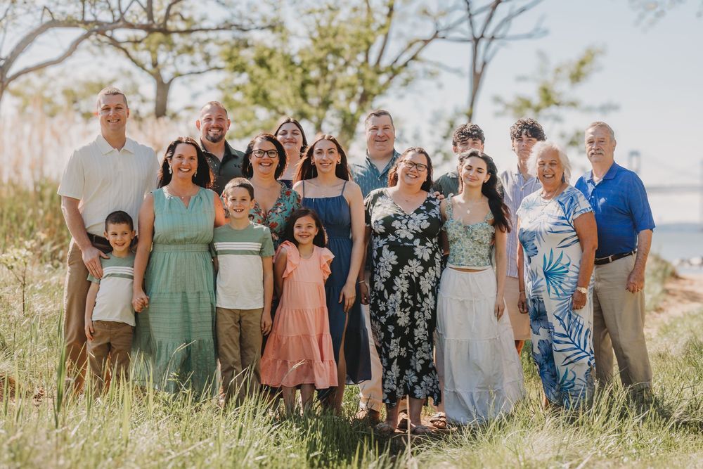 Large family group portrait outdoors in a grassy field near water on a sunny day.