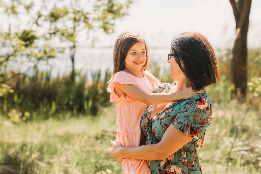 A woman in a floral dress holds a smiling girl in a pink dress outdoors in a sunny green field.