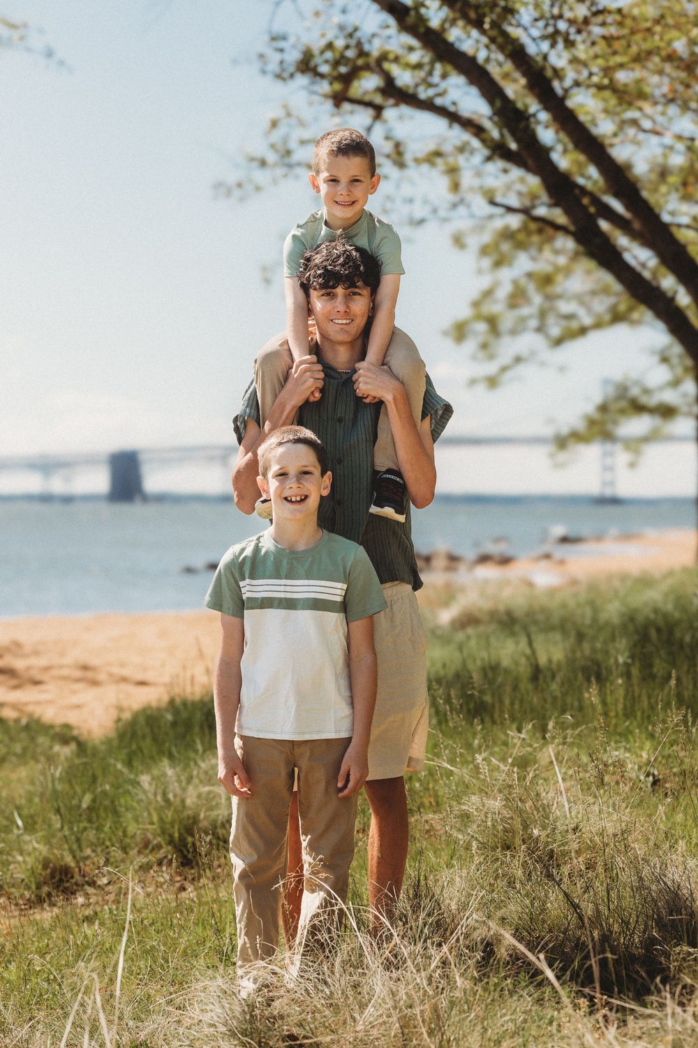 A man carries a child on his shoulders while another boy stands in front, posing near a sunny beach with a bridge in the background.