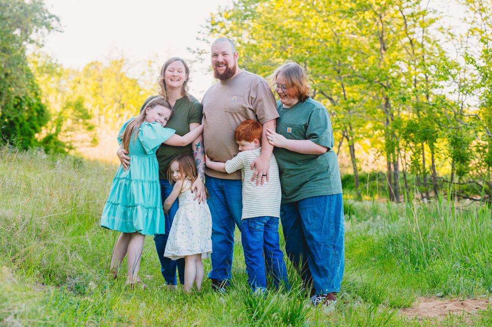 Family of five smiling and embracing outdoors in a green meadow with trees during golden hour.