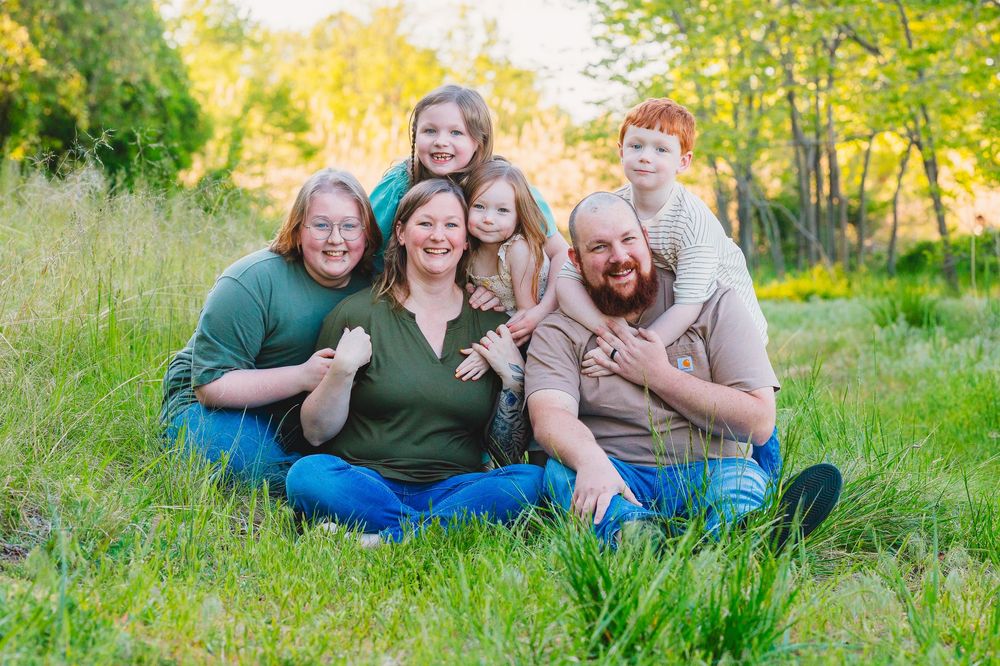 Happy family of six smiling together on green grass outdoors, surrounded by lush trees in warm golden sunlight.