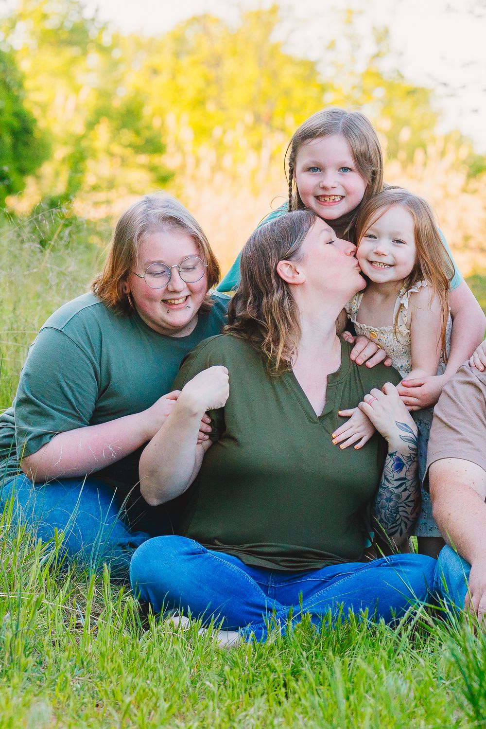 A mother sits in green grass kissing her young daughter while two other girls smile and cuddle nearby outdoors.