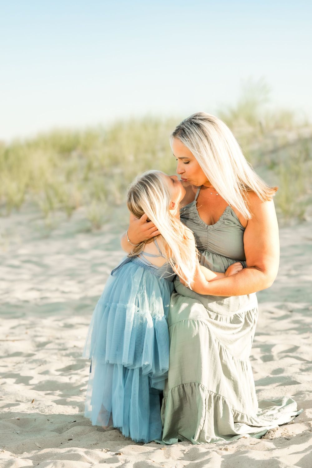 A blonde mother kisses her young daughter on a sandy beach, both wearing soft sage and blue dresses.