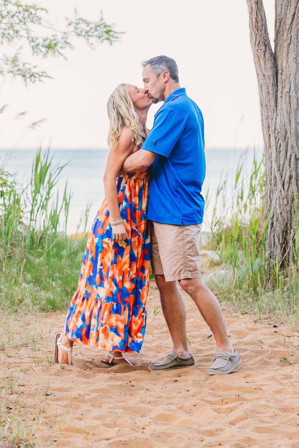 Couple kissing on sandy beach path, woman in colorful blue and orange floral maxi dress, man in blue polo and khaki shorts.