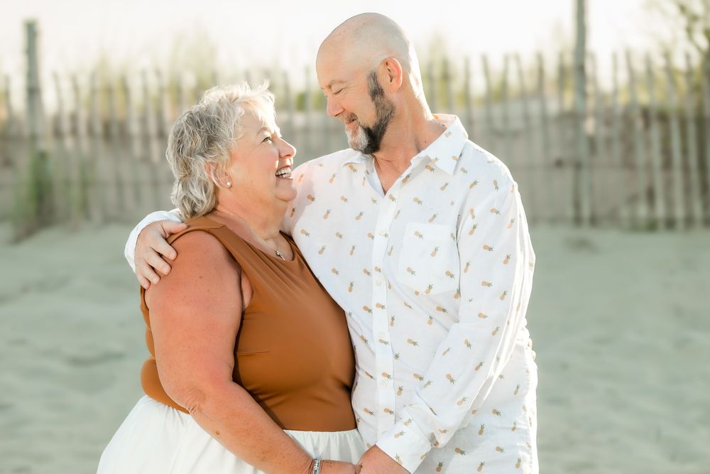Elderly woman and bald man smiling at each other on a sandy beach, sharing a warm embrace near a wooden fence.