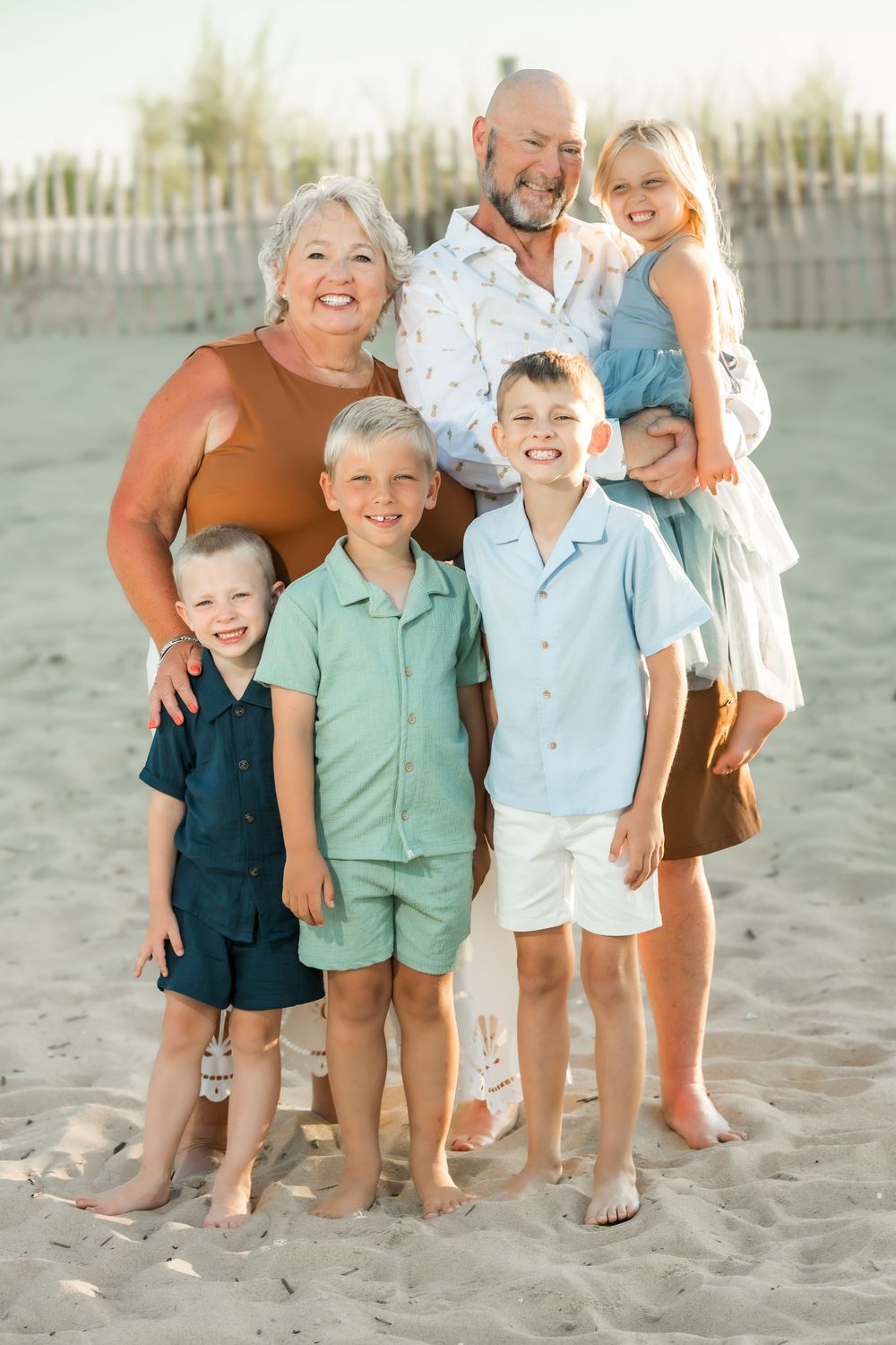 Grandparents and four young grandchildren pose together smiling on a sandy beach during a family photo session.