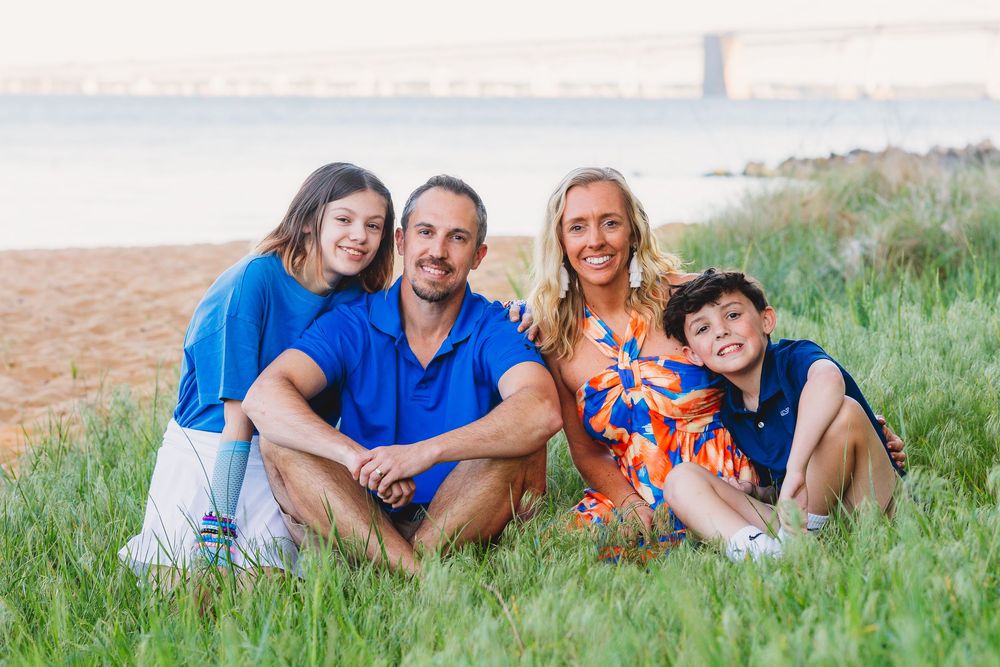 Family of four smiling together on a grassy beach with a bridge in the background on a sunny day.