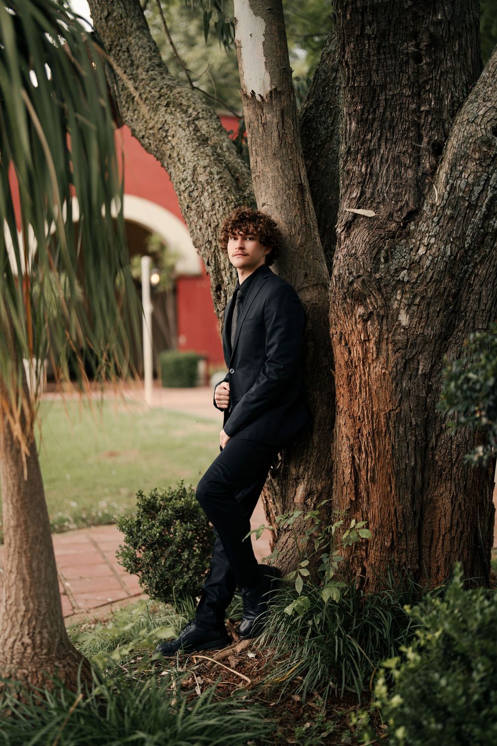 A stylish figure in black attire poses against a tree trunk with a red building in the background.