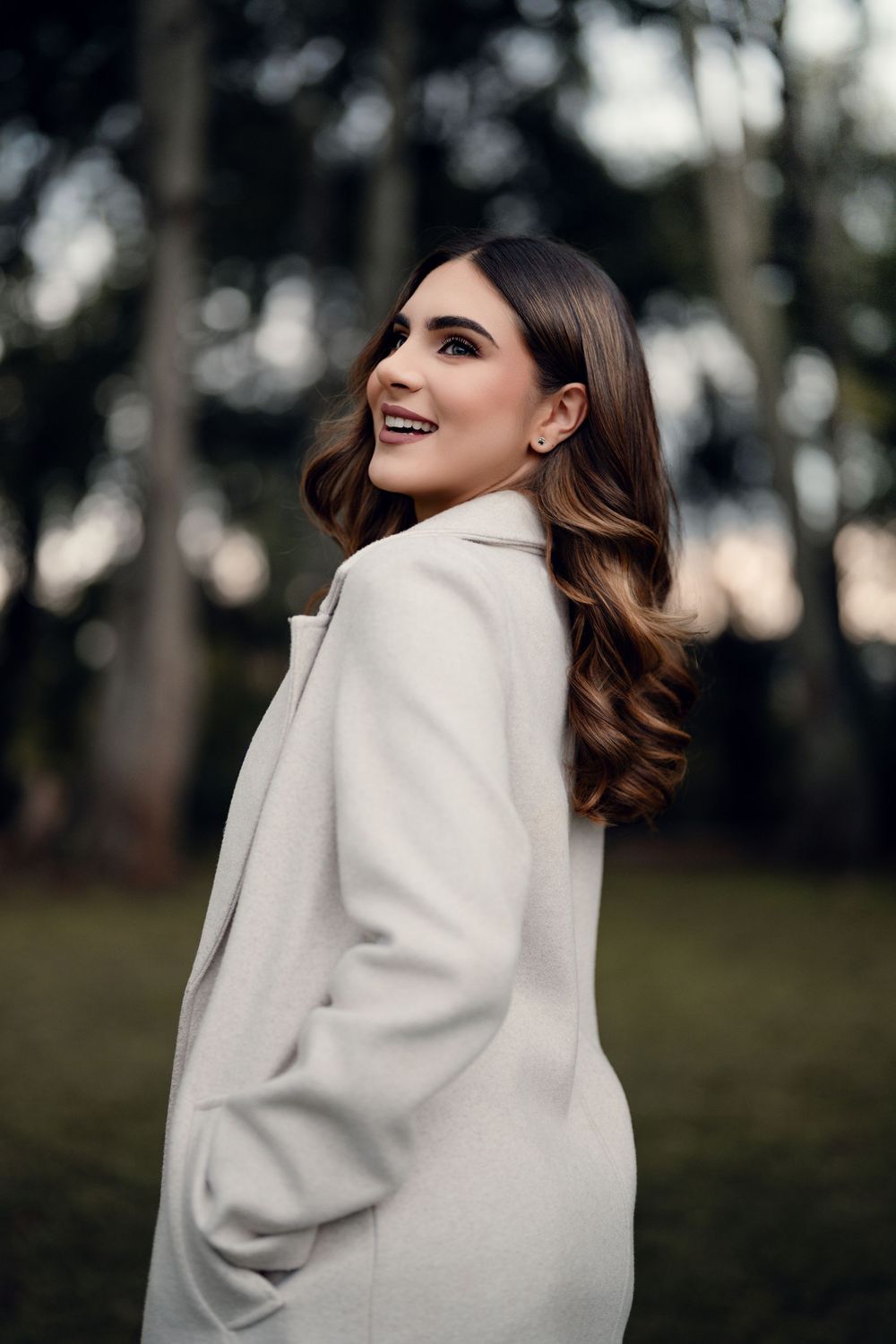 Elegant portrait featuring a cream wool coat and styled waves against a forest backdrop.