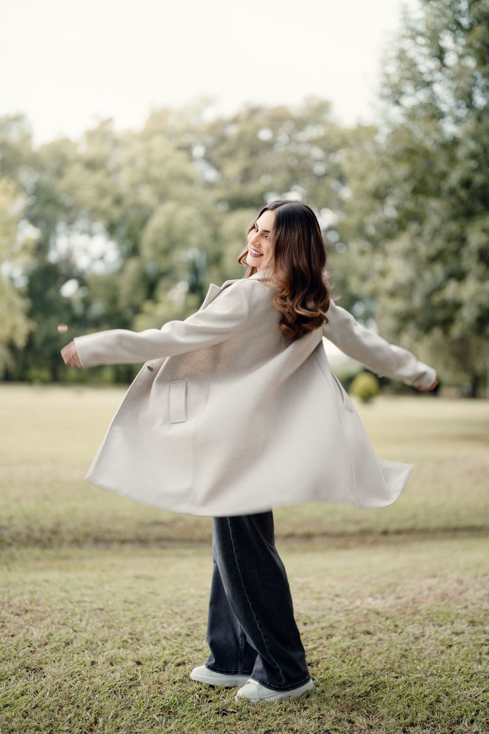 Joyful twirling motion in a cream coat and wide-leg jeans on a grassy lawn.