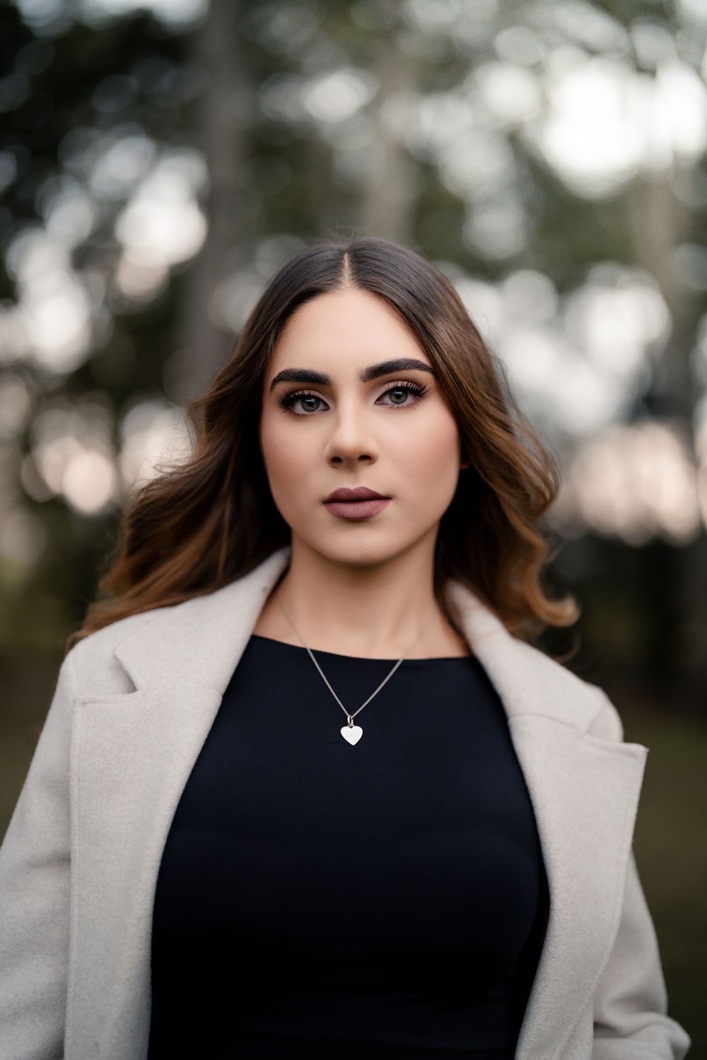 Portrait featuring a cream coat, black top, and delicate heart necklace against a blurred natural backdrop.