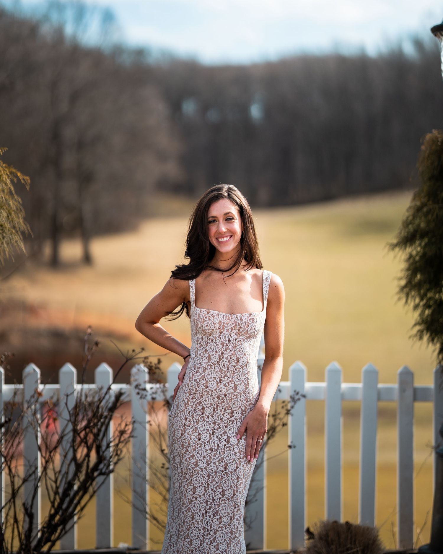A woman in a lace dress poses by a white picket fence in a countryside setting.