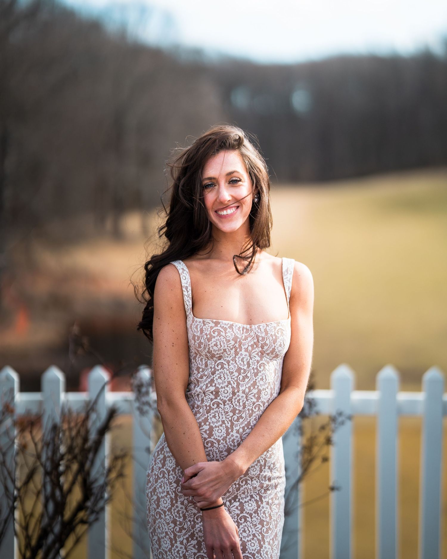 A portrait of a woman in a lace dress standing by a white picket fence.