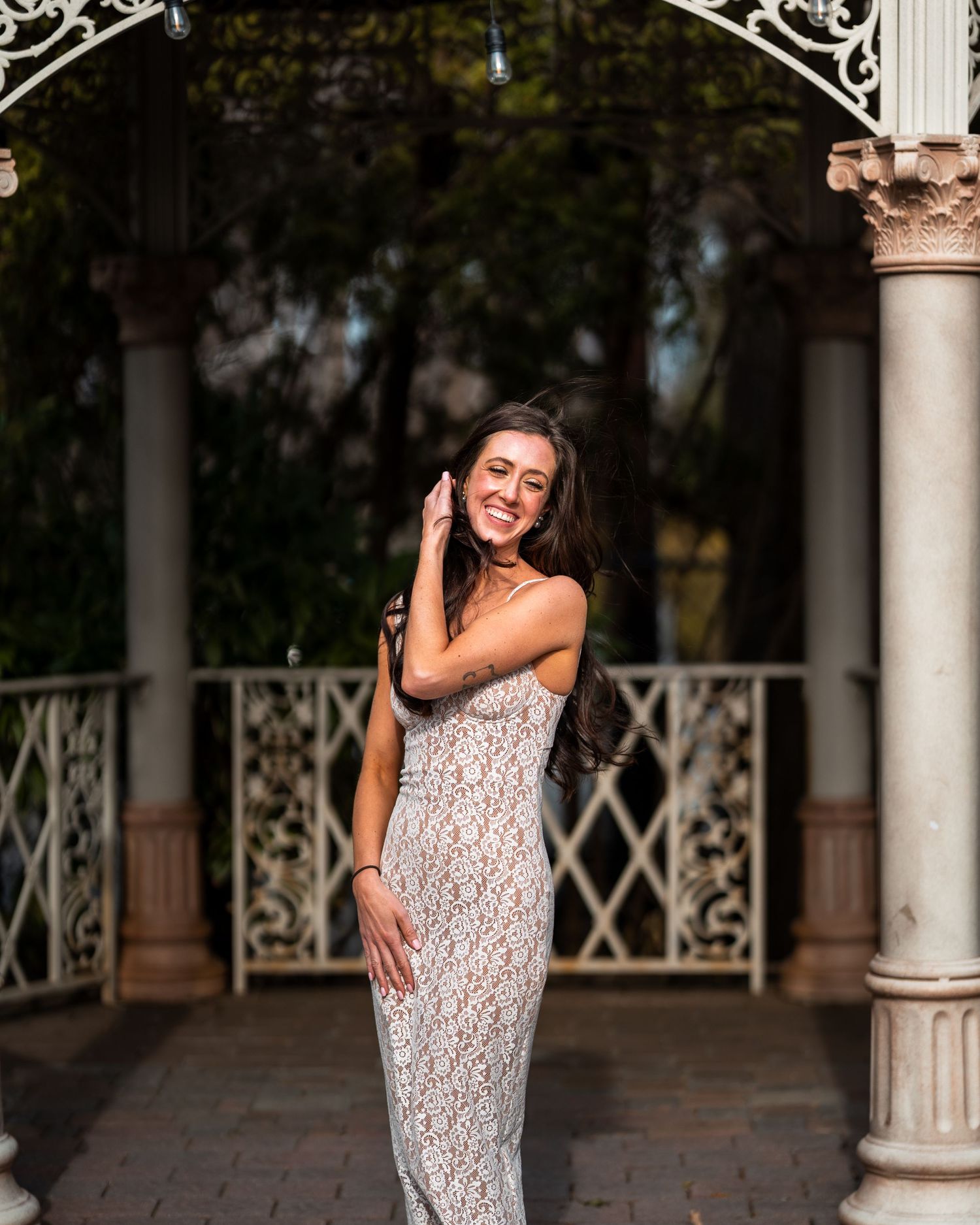 A woman in a lace dress poses playfully under an ornate garden gazebo.