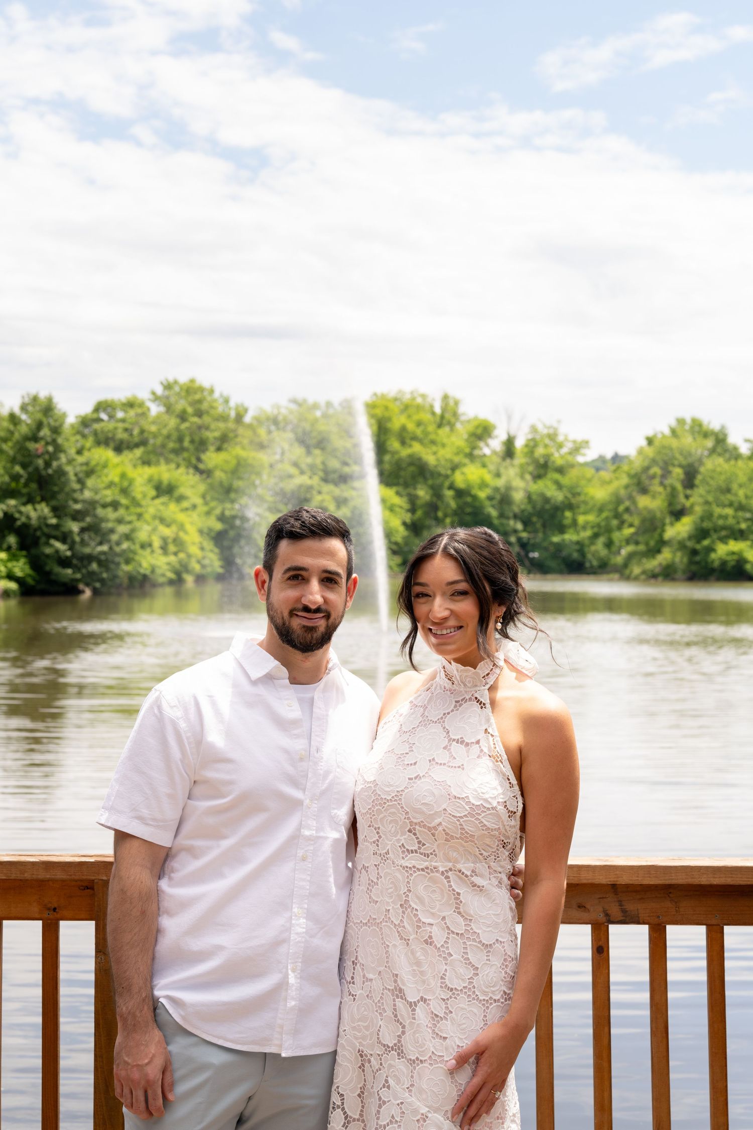 A couple poses together on a deck overlooking a lake with a fountain in the background.
