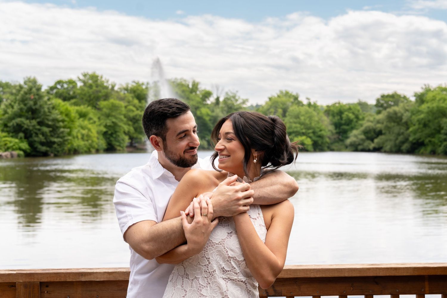 A couple embraces and laughs together on a wooden deck by a scenic lake.