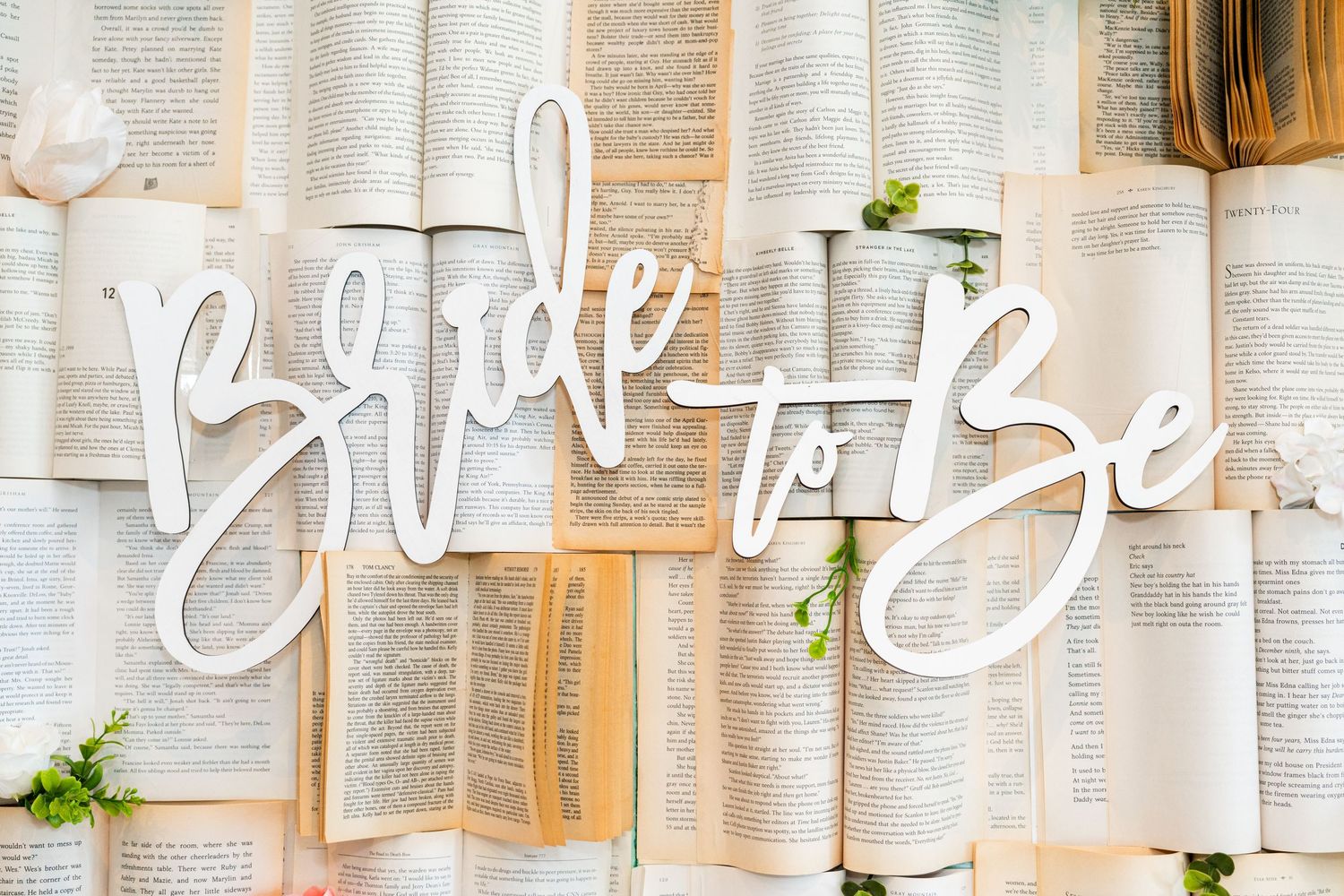 White 'Bride to Be' text displayed on a creative wall made of open book pages with greenery accents.