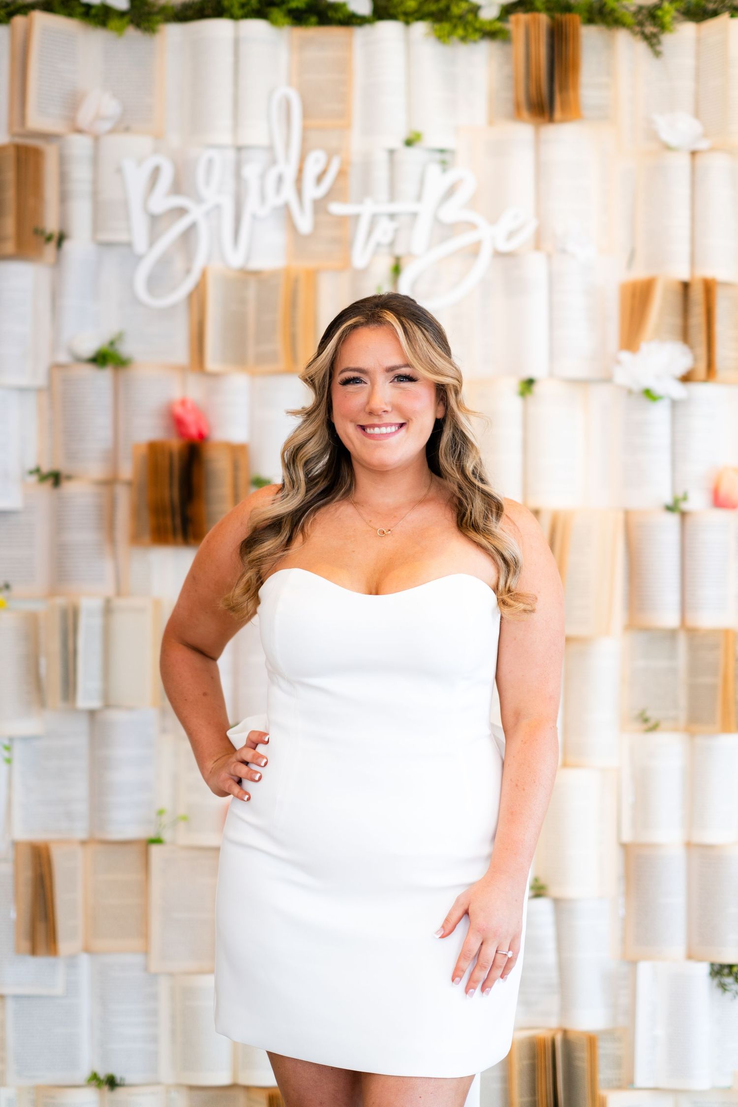 A woman in a white dress poses in front of a decorative backdrop with 'Bride to Be' signage.