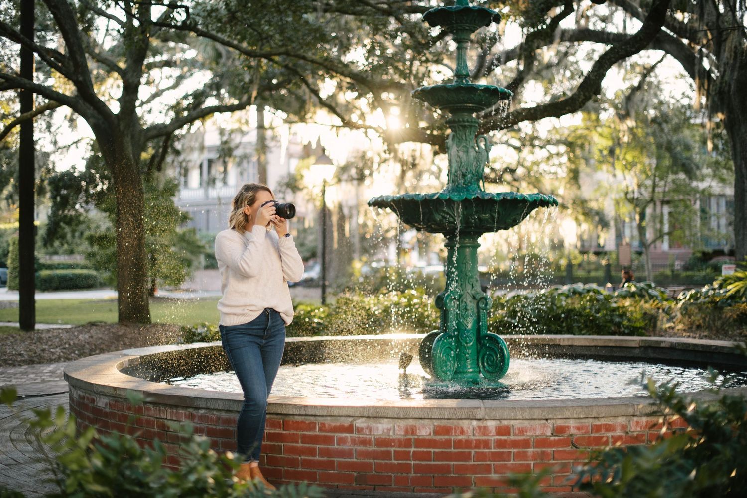 A vintage green fountain stands in a brick-lined courtyard at golden hour with trees in the background.