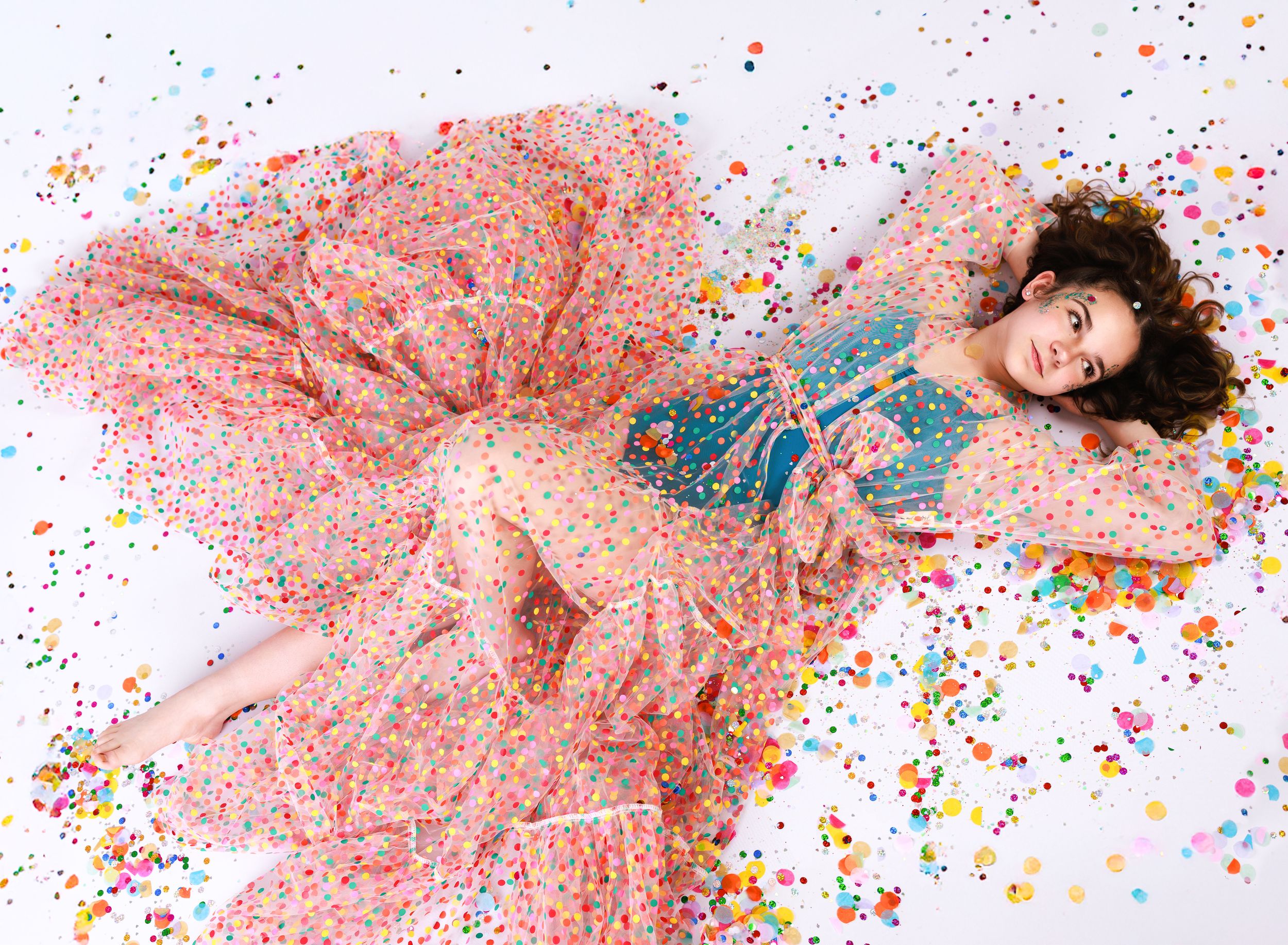 A dreamy portrait on a floor surrounded by colorful confetti with a flowing pink polka dot dress.