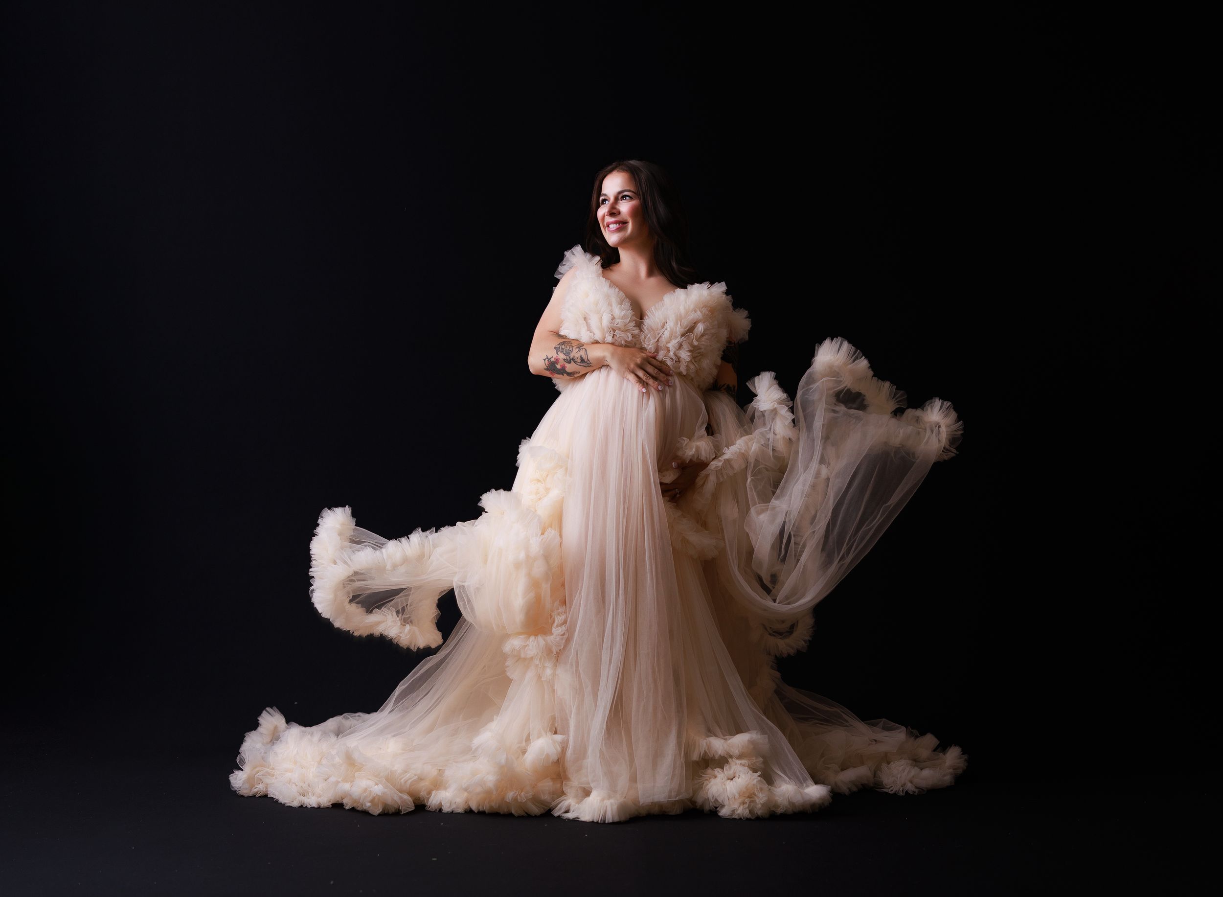 A figure in a flowing blush tulle gown poses dramatically against a dark background in an elegant maternity portrait.