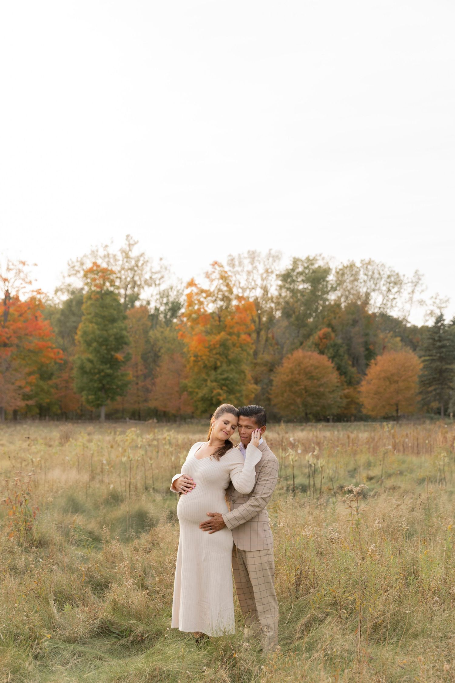 man in a tan suit holding his pregnant wife's belly as they cuddle up close together in a field in the autumn