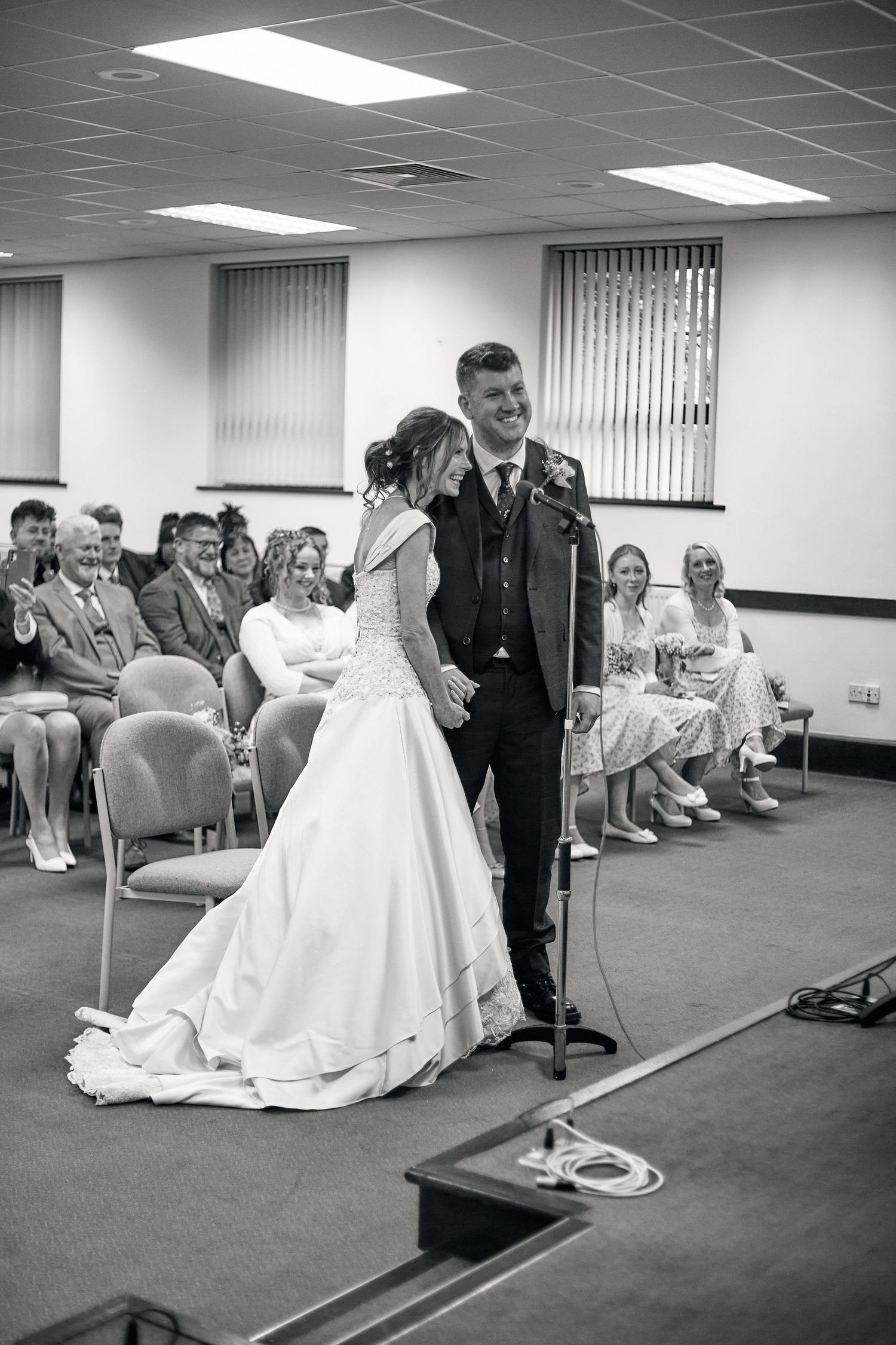 A bride and groom stand at a microphone during their wedding ceremony with guests seated in rows of chairs.