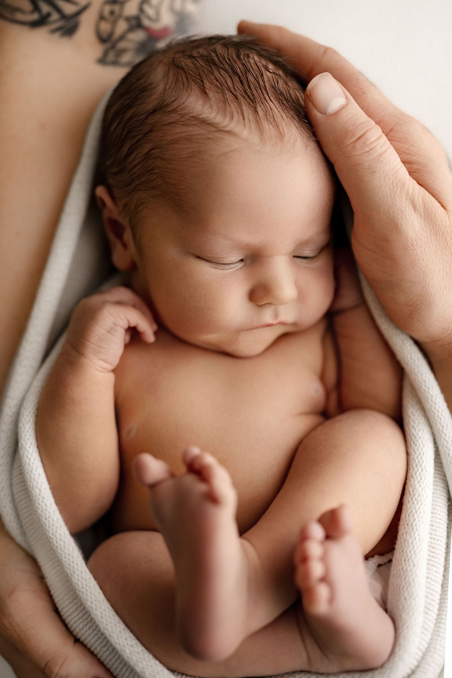 Newborn baby wrapped in white fabric held gently by tattooed hands.