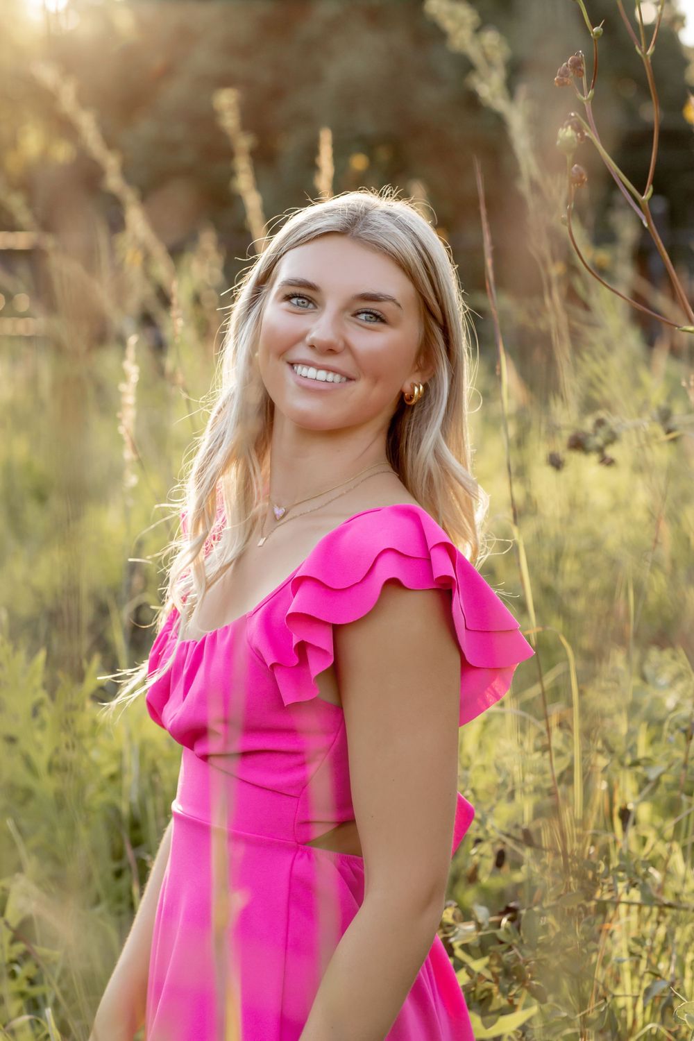 Smiling blonde woman in pink ruffled dress standing in golden sunlit field.