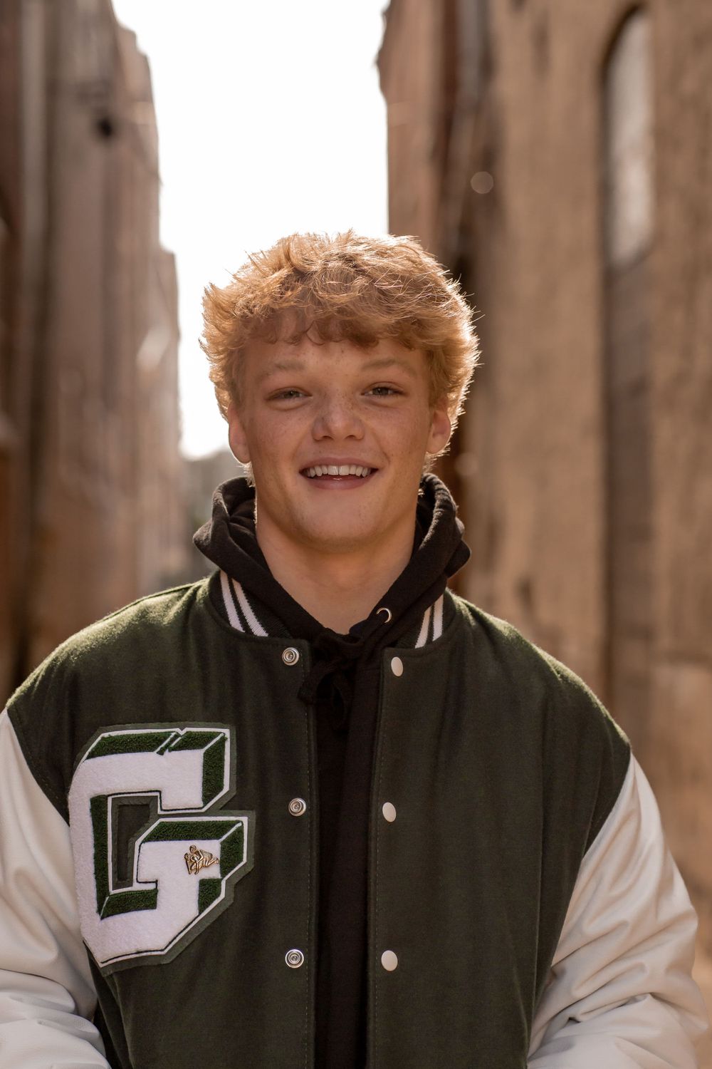 A smiling teen boy in a green and white varsity jacket poses in an urban alleyway.