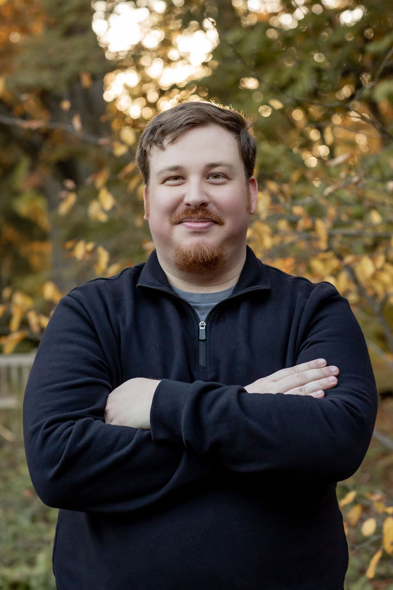 A bearded man in a black zip-up sweater stands with arms crossed smiling outdoors in fall.
