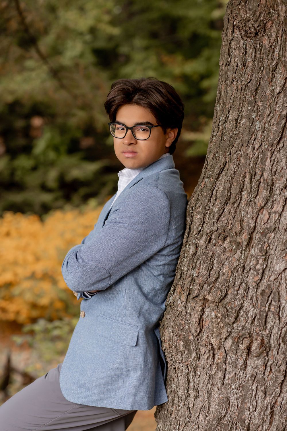 A young man in a grey blazer and glasses leans against a tree trunk in an autumn park.