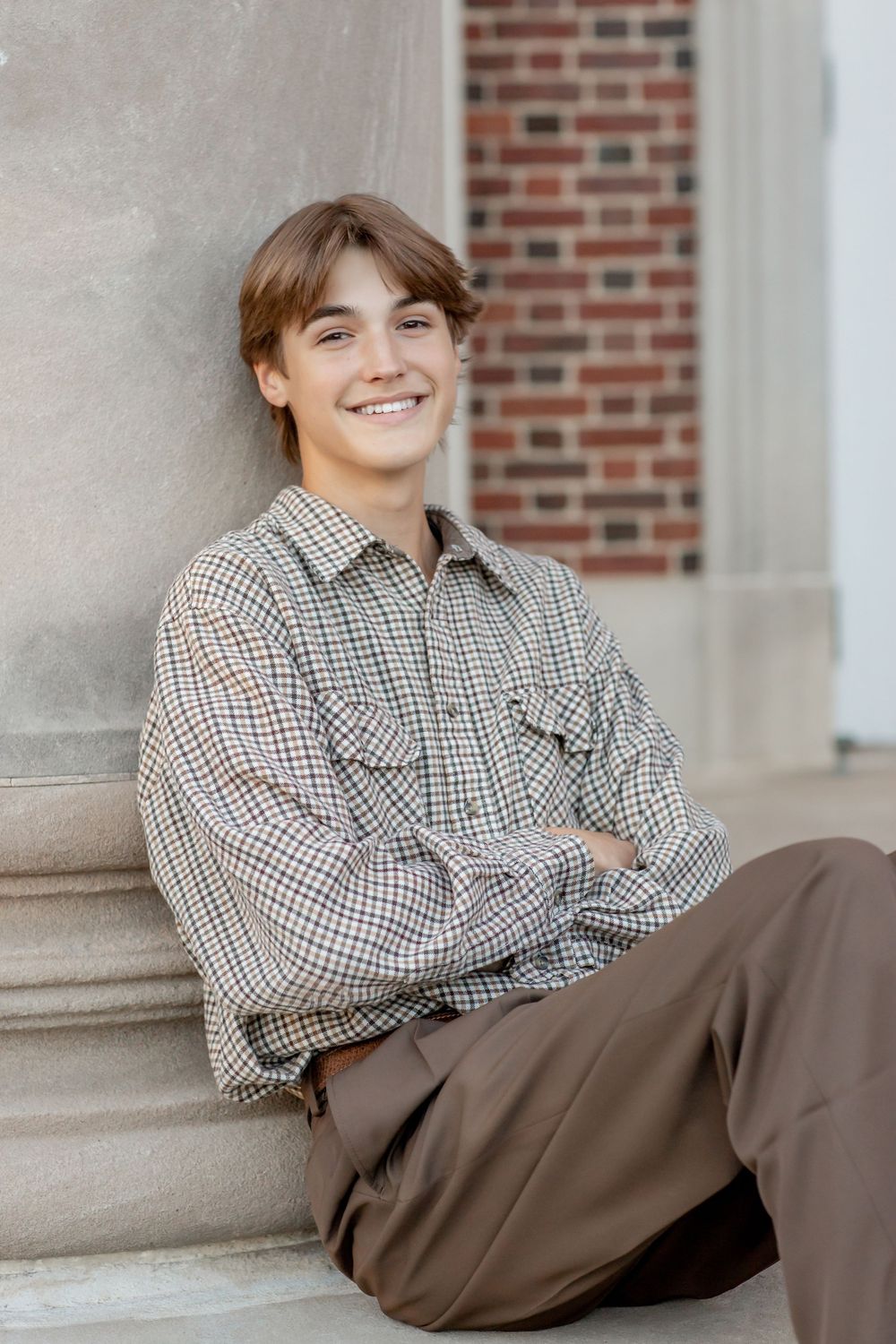 Smiling teenage boy in plaid shirt leaning against stone column outdoors.